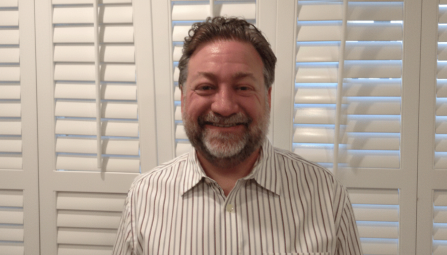 Man with a beard and mustache wearing a striped shirt standing in front of white window blinds.