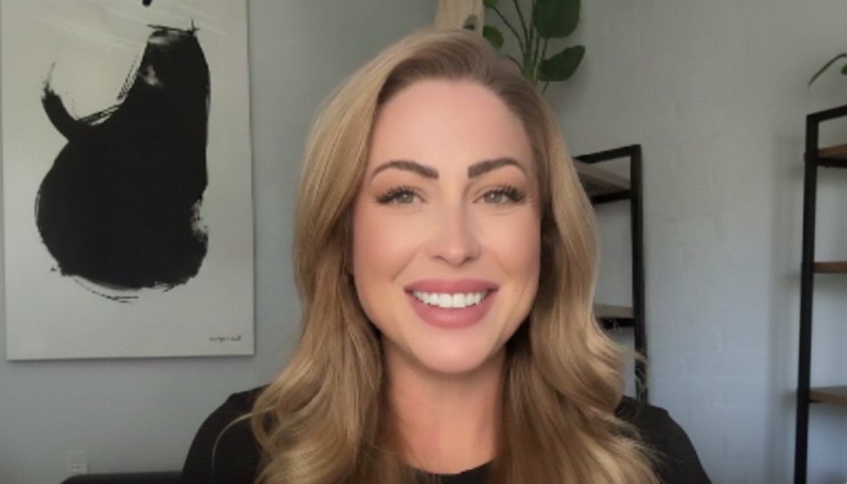 Woman with long blonde hair smiling in a room with a painting and shelves in the background